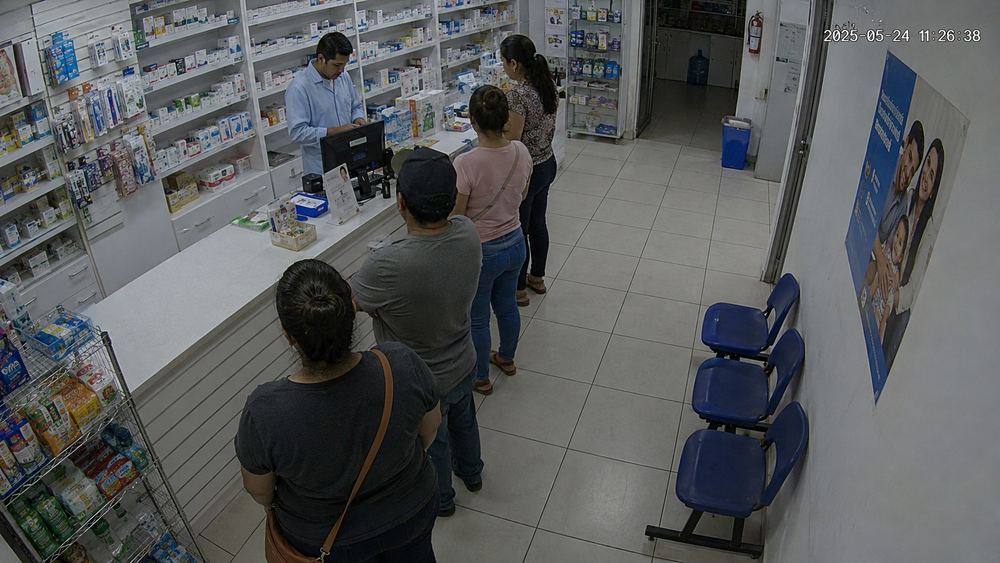 Cámara cenital de una farmacia: cuatro clientes formados en diagonal hacia el mostrador, y un empleado atendiendo detrás.