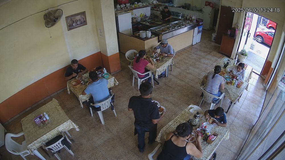 Cámara cenital de una fonda a la hora de la comida: cuatro mesas ocupadas, una libre, y un mesero llevando platos.
