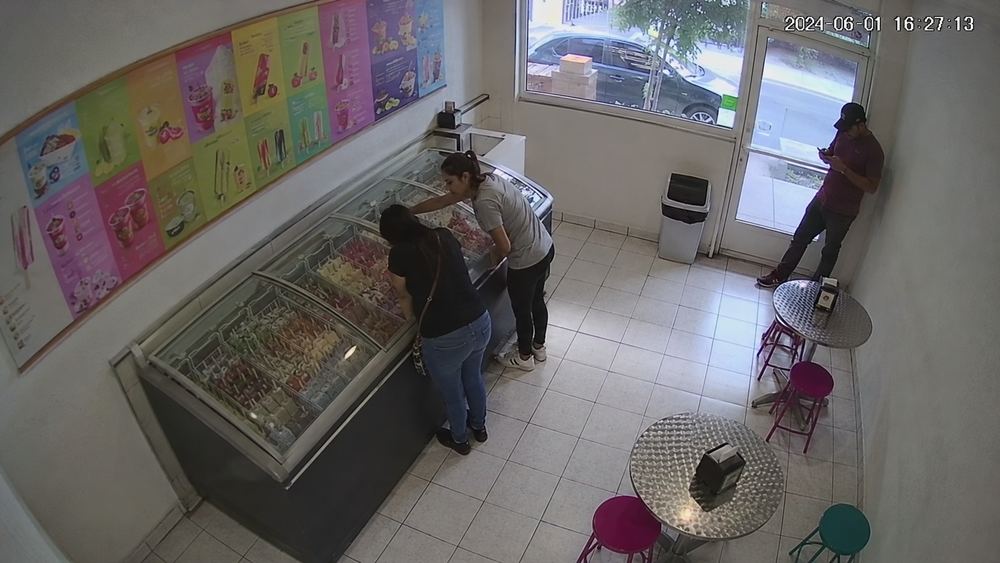 Cámara cenital de una paletería: dos personas en la vitrina de helados y una junto a la puerta; las mesas del fondo están vacías.